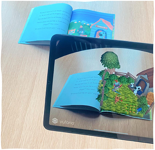 An open children's book lies on a table, and a tablet displays an augmented reality scene from the book. The scene shows a 3D farmyard with a tree, a doghouse, and a dog. The book features bright illustrations.