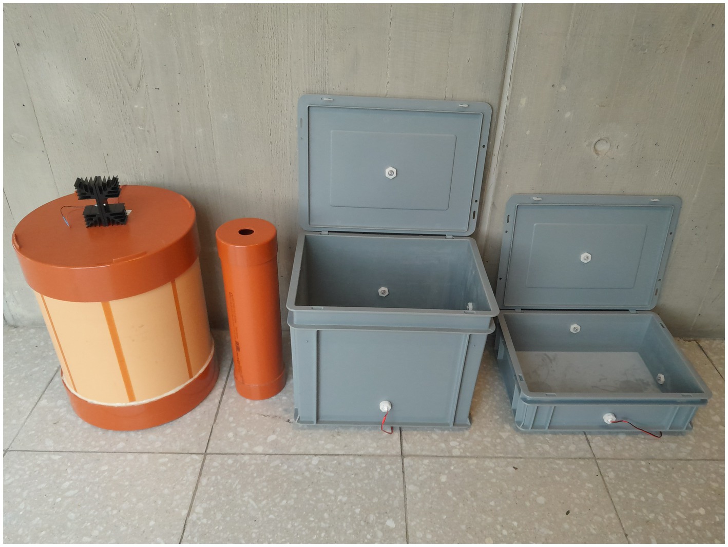 Various equipment pieces are displayed against a concrete wall and on a tiled floor. They include a large and a small cylindrical orange with the same height but different diameters. The large cylindrical container has a heatsink in its lid. Next to the cylindrical containers are two gray rectangular boxes with open lids, showing wiring inside.