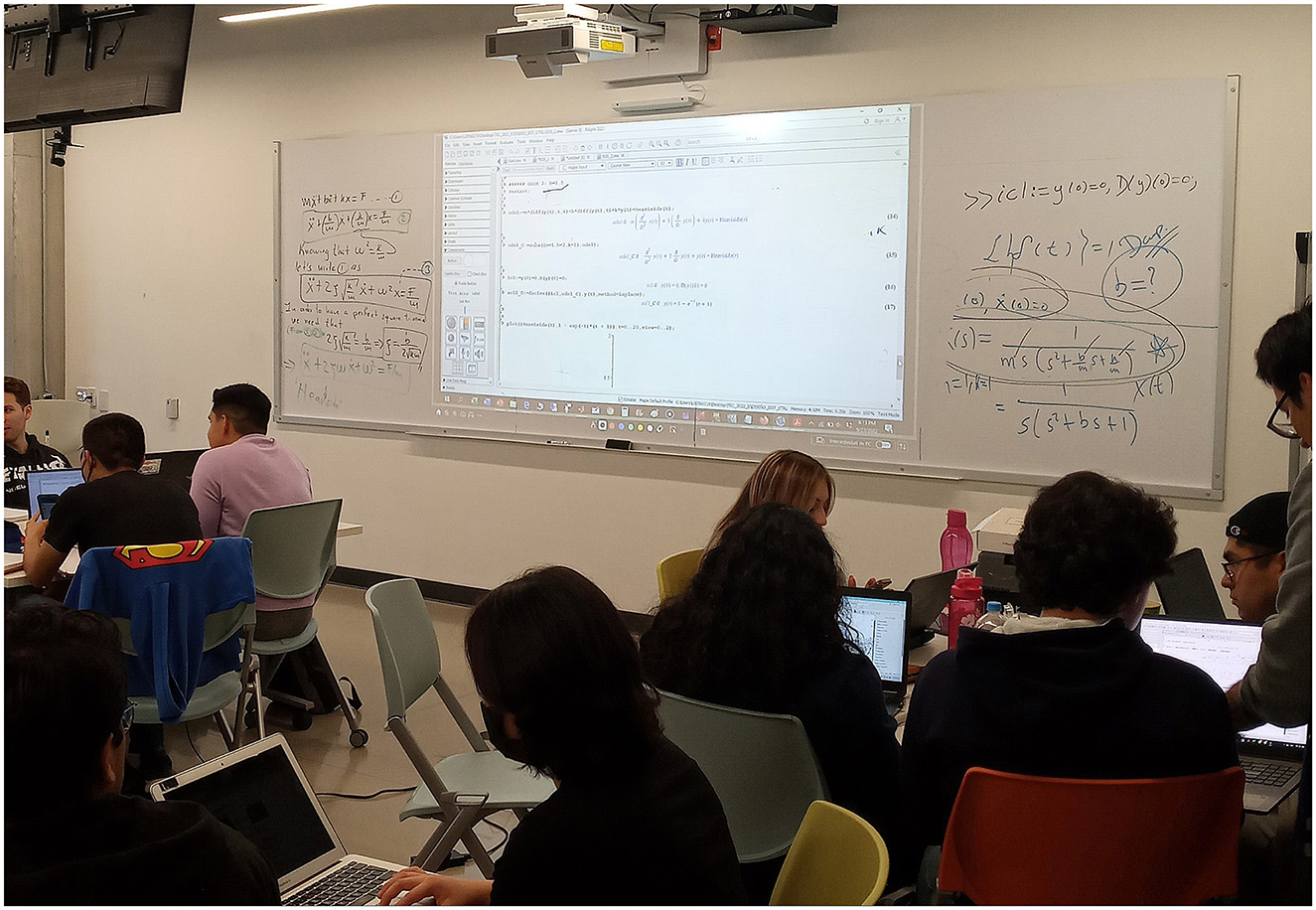 Students are sitting in a classroom in front of a whiteboard displaying complex mathematical equations and a projected computer screen. Some are working on laptops, and one person is wearing a shirt with a Superman logo draped over their chair.