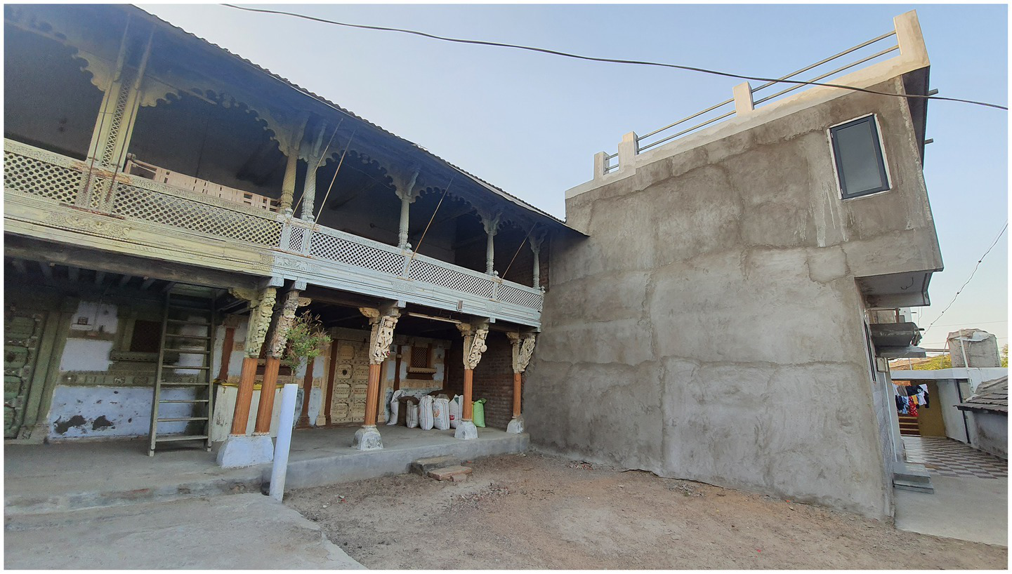 A photograph depicting the hybridisation of the vernacular façade 1, which is a two-story building with exquisite wooden carvings and an open balcony supported by decorative pillars standing next to a simple, concrete structural extension. The ground is rough and uneven, with a staircase linking the floors on the veranda and numerous sacks piled in the corner.