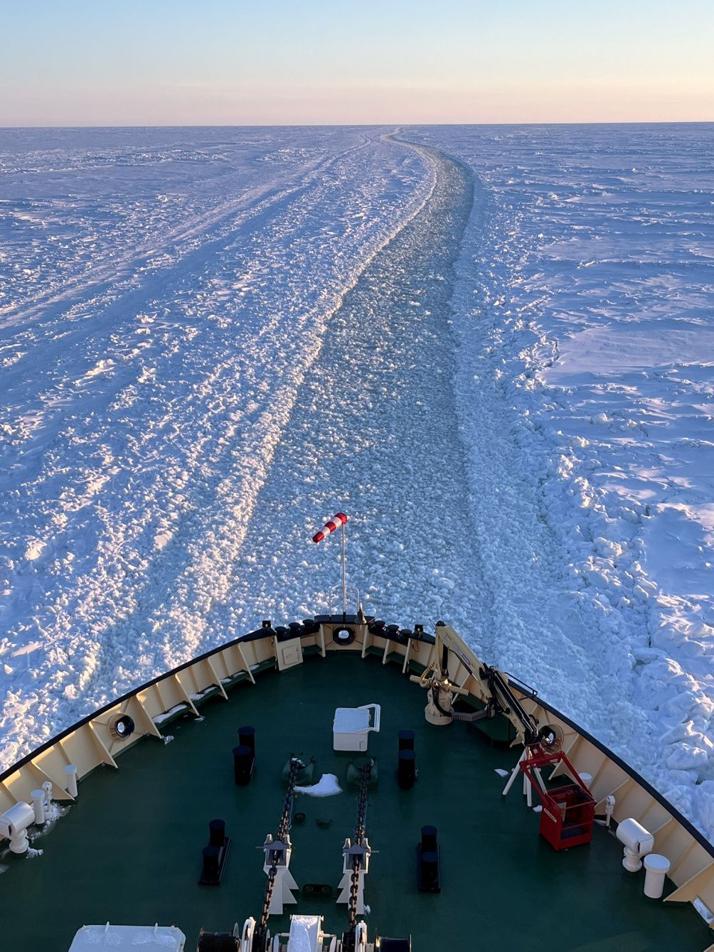 An ice field with a ship track in the middle with pieces of broken ice and a ship's bow is seen in the track.