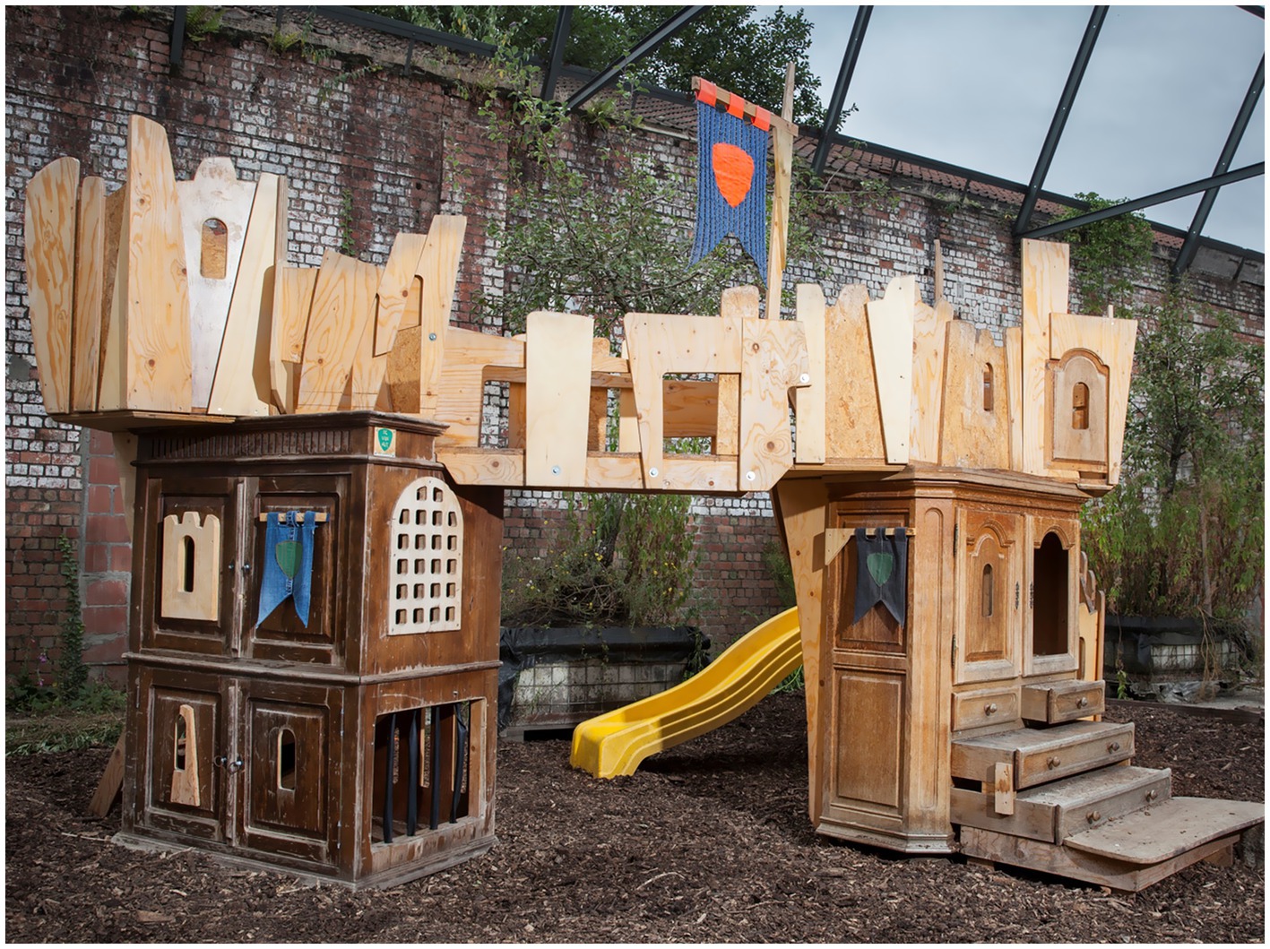 A wooden playground structure designed to look like a castle. It features two upcycled big wardrobes turned into towers and connected by a bridge, and it has colourful banners and a slide. It was made for the public garden of a large cohousing community by one of the makers in the cooperative.