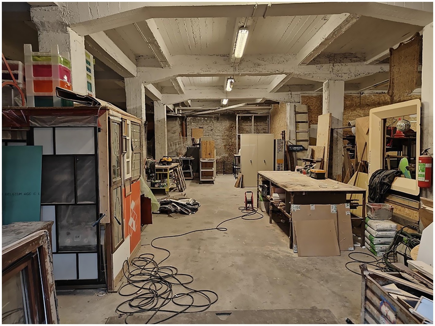 Interior of a workshop with exposed walls and ceilings and industrial lighting. The space is cluttered with workbenches, tools, and materials. Wooden panels, cables, and equipment are scattered throughout. This is the tool-sharing space of the cooperative.