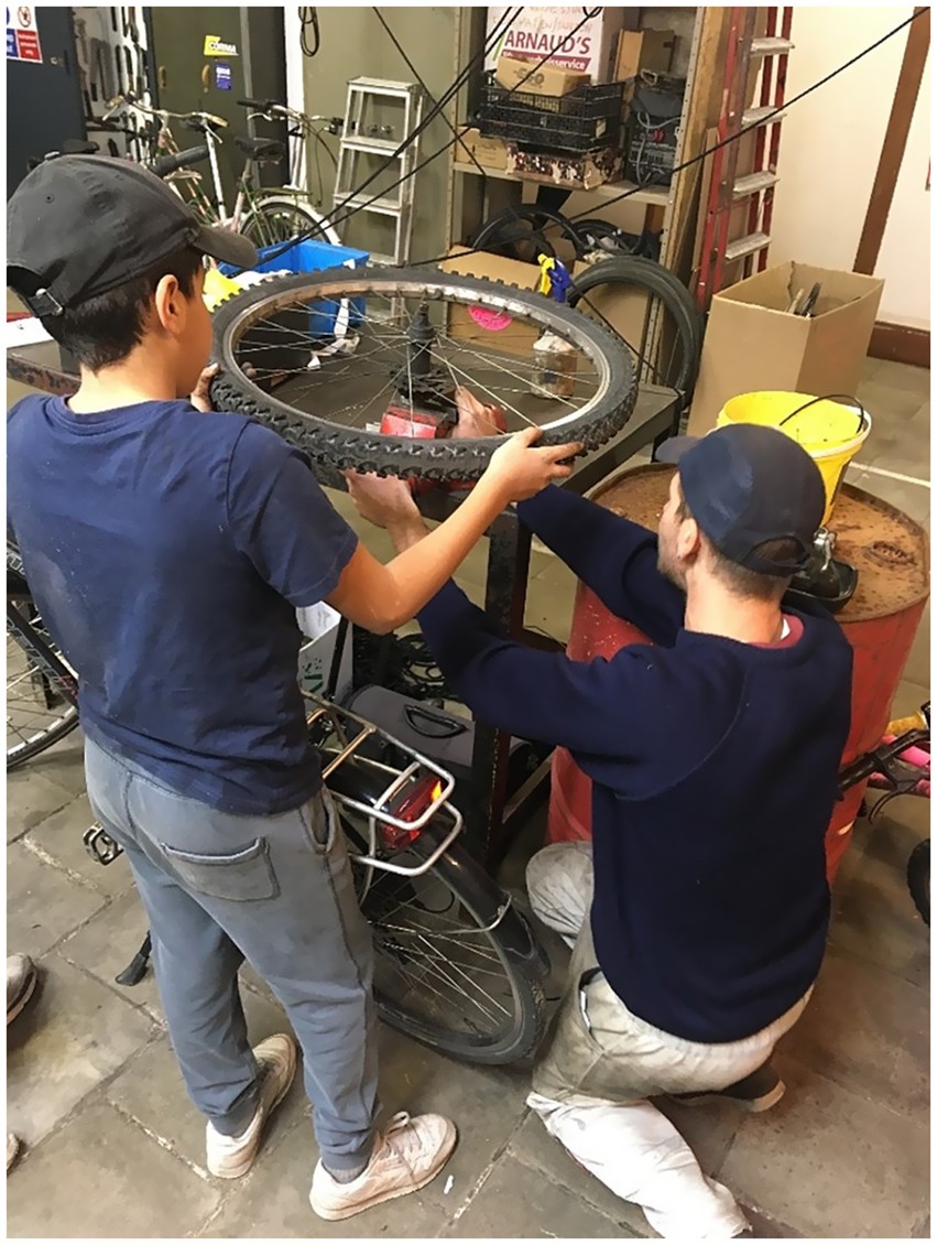 Two people, a child and the teacher-mechanic, work together in a bike repair shop. The child, who is being taught, holds a bicycle wheel, while the teacher kneels and makes a repair intervention. Various bike parts and tools surround them in the workshop.
