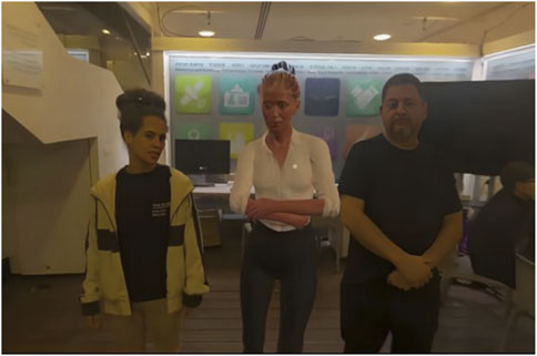 Three people are standing indoors. The individual on the left wears a light jacket over a black shirt. The middle figure appears to be a robot with a human-like appearance, wearing a white shirt. The person on the right is dressed in a black shirt. There is a computer and a colorful wall display in the background.