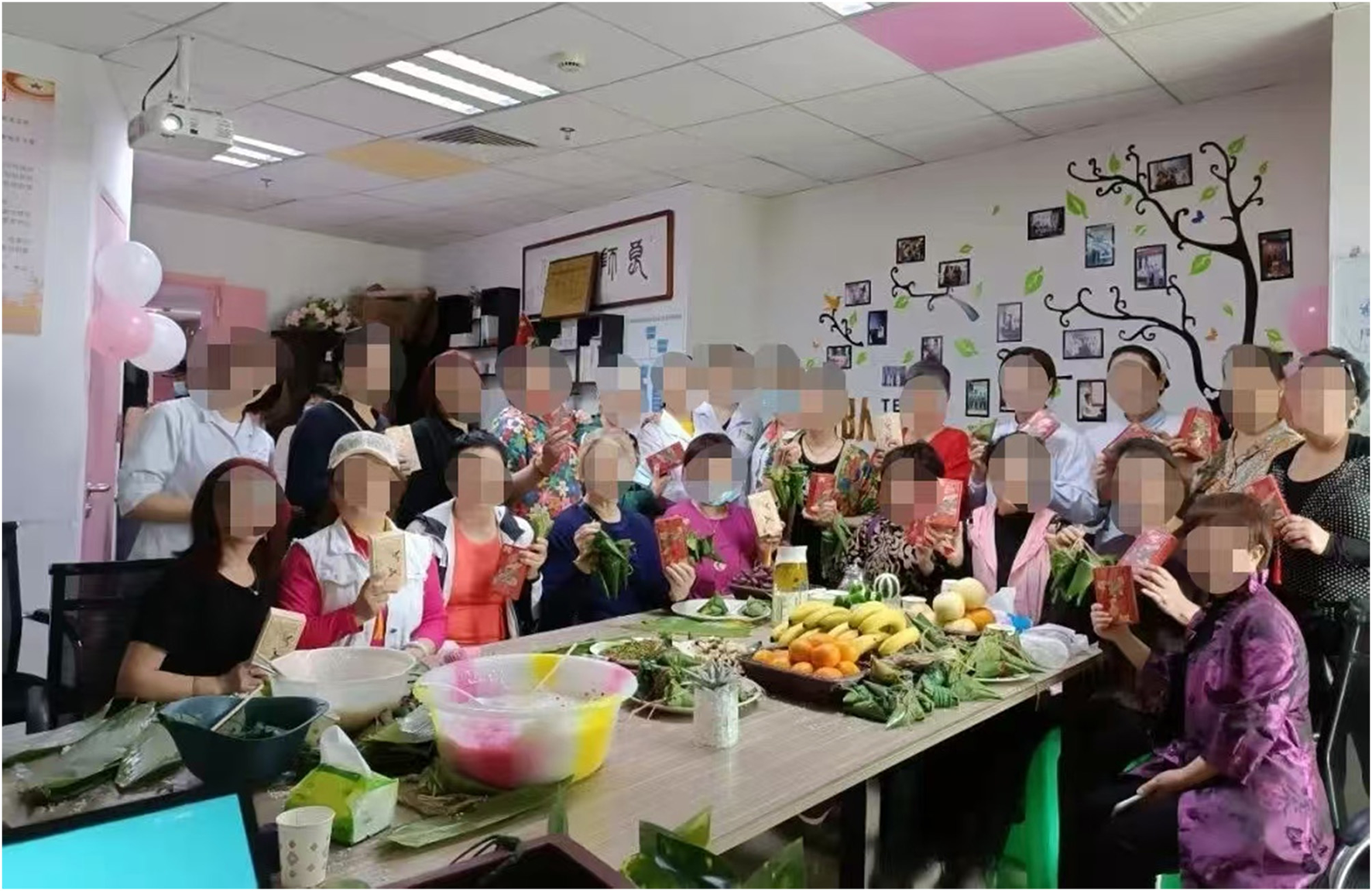 A group of people gathered in a decorated room, holding red envelopes and Zongzi, a traditional Chinese food made of rice wrapped in leaves. A table in front has fruit, snacks, and materials for making Zongzi. The wall is adorned with framed photos and a painted tree design