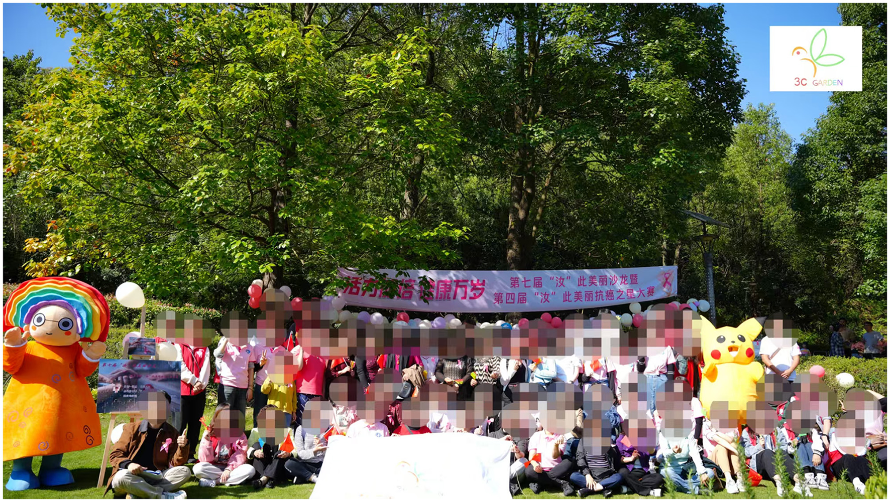 A large group of people, including children, pose outdoors in front of a banner and colorful balloons. The setting is a grassy area with trees. There are two large character costumes, one resembling a yellow creature and the other with a rainbow head, on either side of the group.