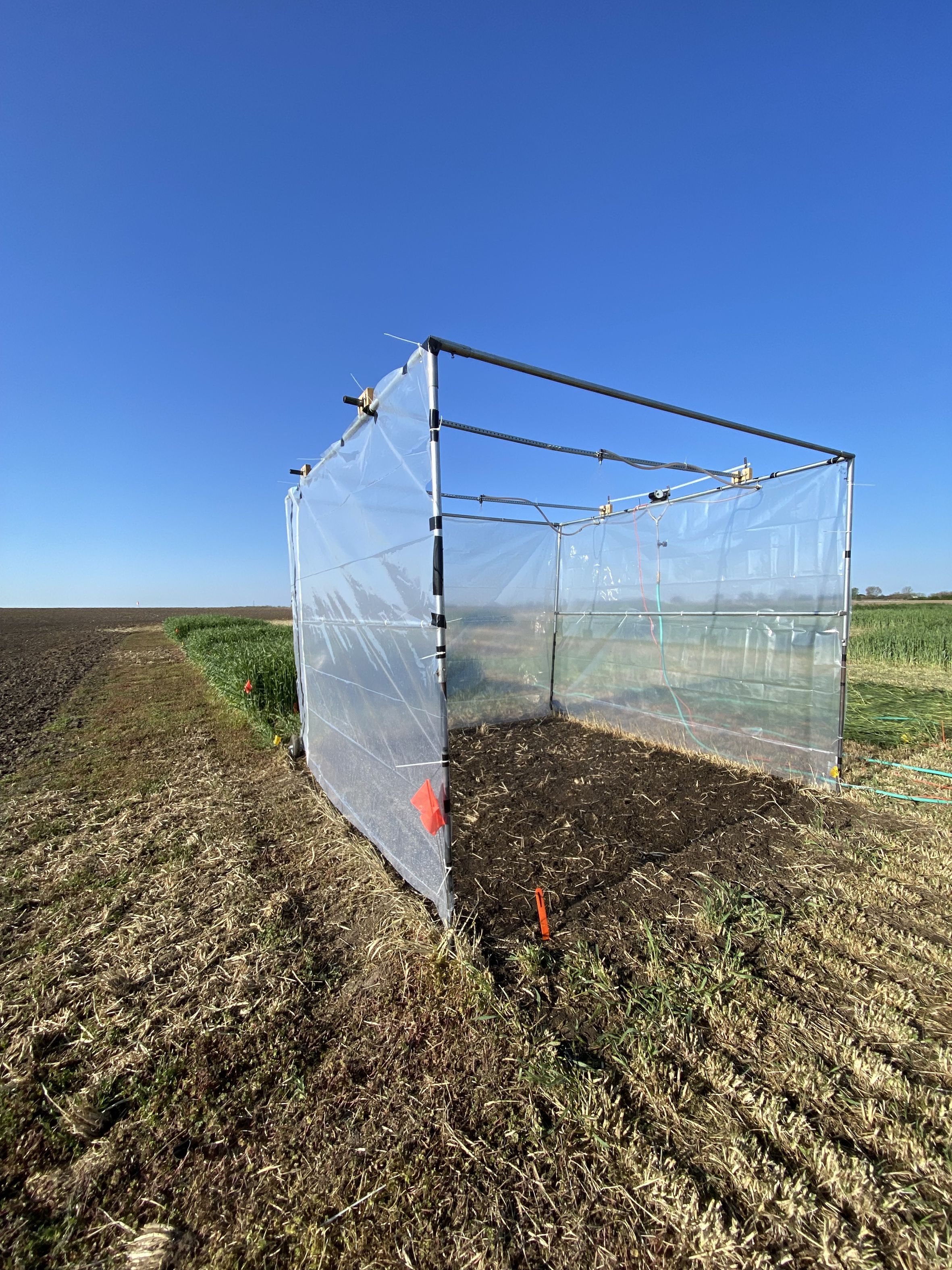 A transparent plastic enclosure stands on a grassy field under a clear blue sky. The structure is supported by a metal frame with visible joints and connectors, and it appears to cover a section of soil. Orange markers are placed around the enclosure.