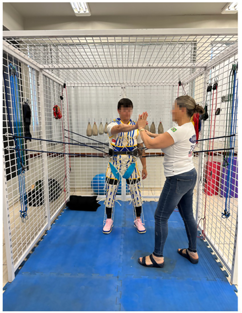 A person is engaged in physical therapy, wearing a harness and supported by elastic cords inside a metal cage structure. They are performing exercises with the assistance of a therapist who is guiding them. Blue mats cover the floor.