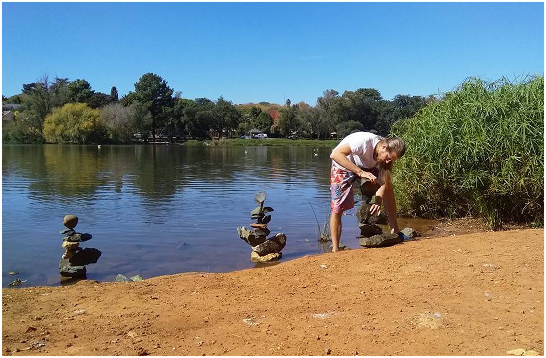 A person balances stones in vertical sculptures by a lake shore on a sunny day. The lake reflects surrounding trees and a clear blue sky. Lush greenery borders the lake.