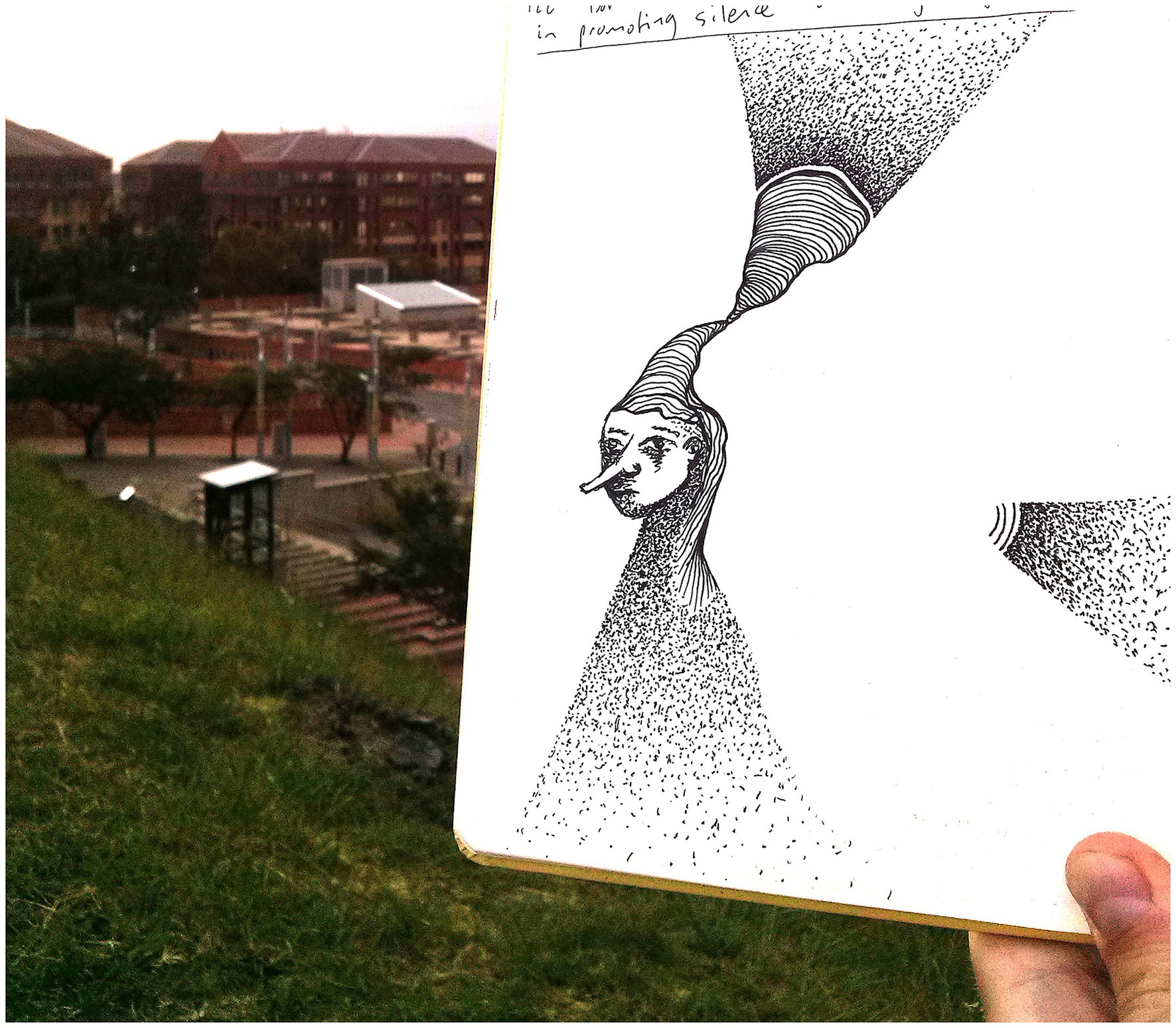 A hand holds an open sketchbook with an abstract drawing of a face with a long nose extending into a flowing textured pattern. The background shows a grassy hill and buildings in the distance.