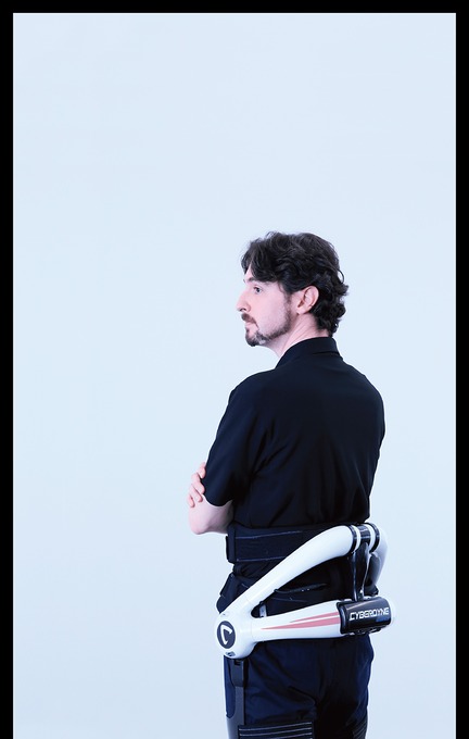 A person wearing a black shirt and a robotic exoskeleton device on their lower back. The device is labeled \