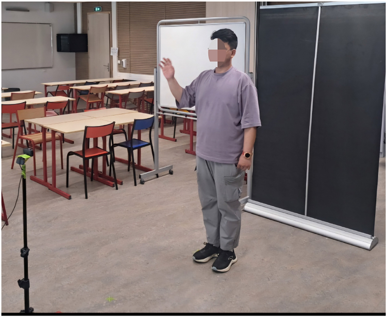 A person stands in a classroom next to a blackboard, raising one hand. Several chairs and desks are arranged in rows, and a digital timer on a stand is visible. The classroom is empty.