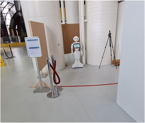 A humanoid robot stands near large cylindrical structures in an indoor setting. It has a screen on its chest displaying an icon. Nearby, there is a tripod and a signboard. Velvet ropes cordon off the area.