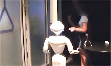 A small humanoid robot is facing a person in a dimly lit room. The person, standing near a table, appears to be interacting with the robot. The setting has a modern, technological appearance.