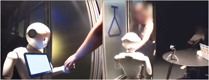 A humanoid robot with a screen on its chest interacts with a person, holding hands. The setting includes a blurred figure, a table, and a tripod with a camera.