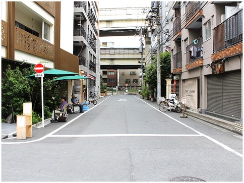 A narrow street in an urban area features tall buildings on both sides. A man sits under a green umbrella on the left sidewalk near a