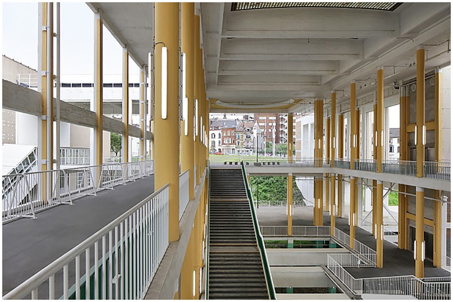 Spacious interior of a modern building featuring multiple levels with open corridors and large yellow columns. Wide staircases connect each level, and bright natural light enters through expansive windows. In the background, there is an urban landscape with buildings.