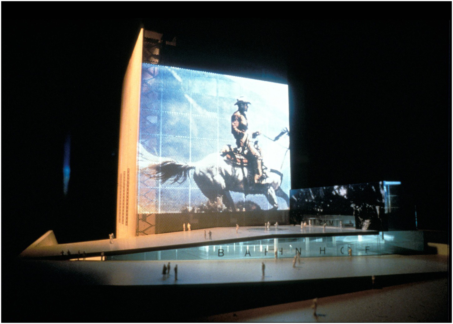 Architectural model featuring a large vertical display with an image of a horse and rider. The structure has a futuristic design with angular elements and small scale figures suggesting a public space.