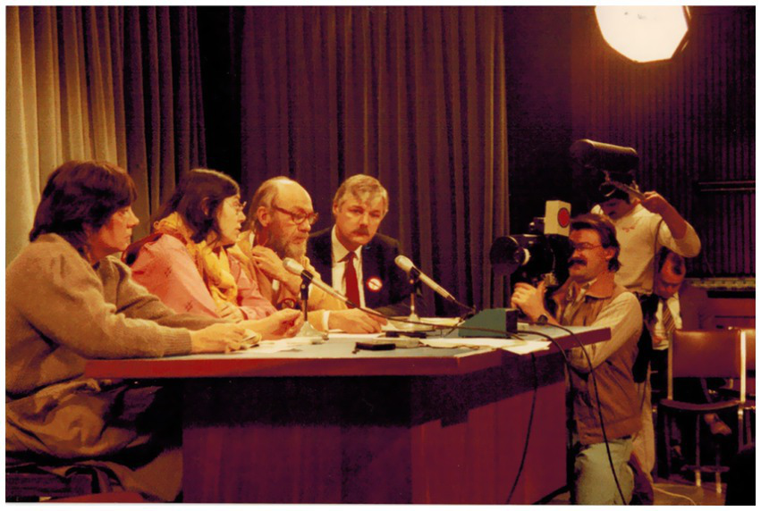 A group of people sit at a table with microphones in front of them, appearing to participate in a panel or discussion. A cameraman is capturing the scene, and another person stands behind him with additional equipment. The setting seems to be indoors with curtains in the background and a bright light overhead.