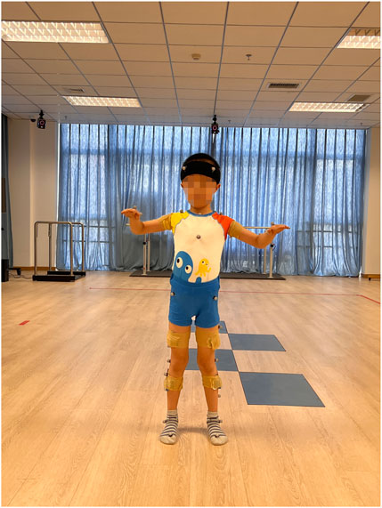 A child standing in a spacious room with wooden floors, wearing a colorful outfit and a helmet. Sensors are attached to their clothing and legs, indicating participation in a research or movement study. The room has blue curtains and several pieces of equipment in the background.