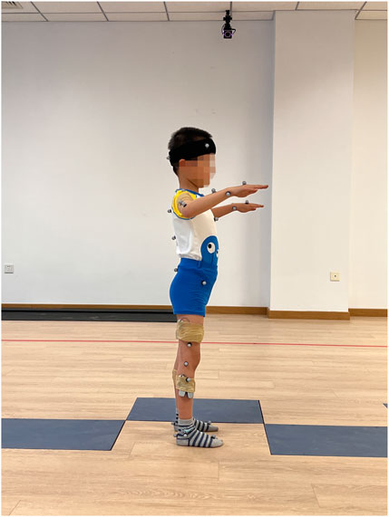 A child facing sideways stands on a wooden floor with arms extended forward. They are wearing blue shorts, a patterned shirt, and striped socks. Motion capture markers are attached to their body, and a headband is worn. The room has blank walls and a camera on the ceiling.