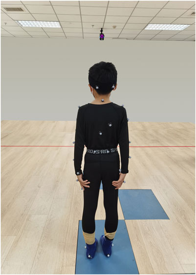 A person stands on a wooden floor wearing a motion capture suit with markers. They face a gray wall with a camera mounted on the ceiling. Blue patches are on the floor.