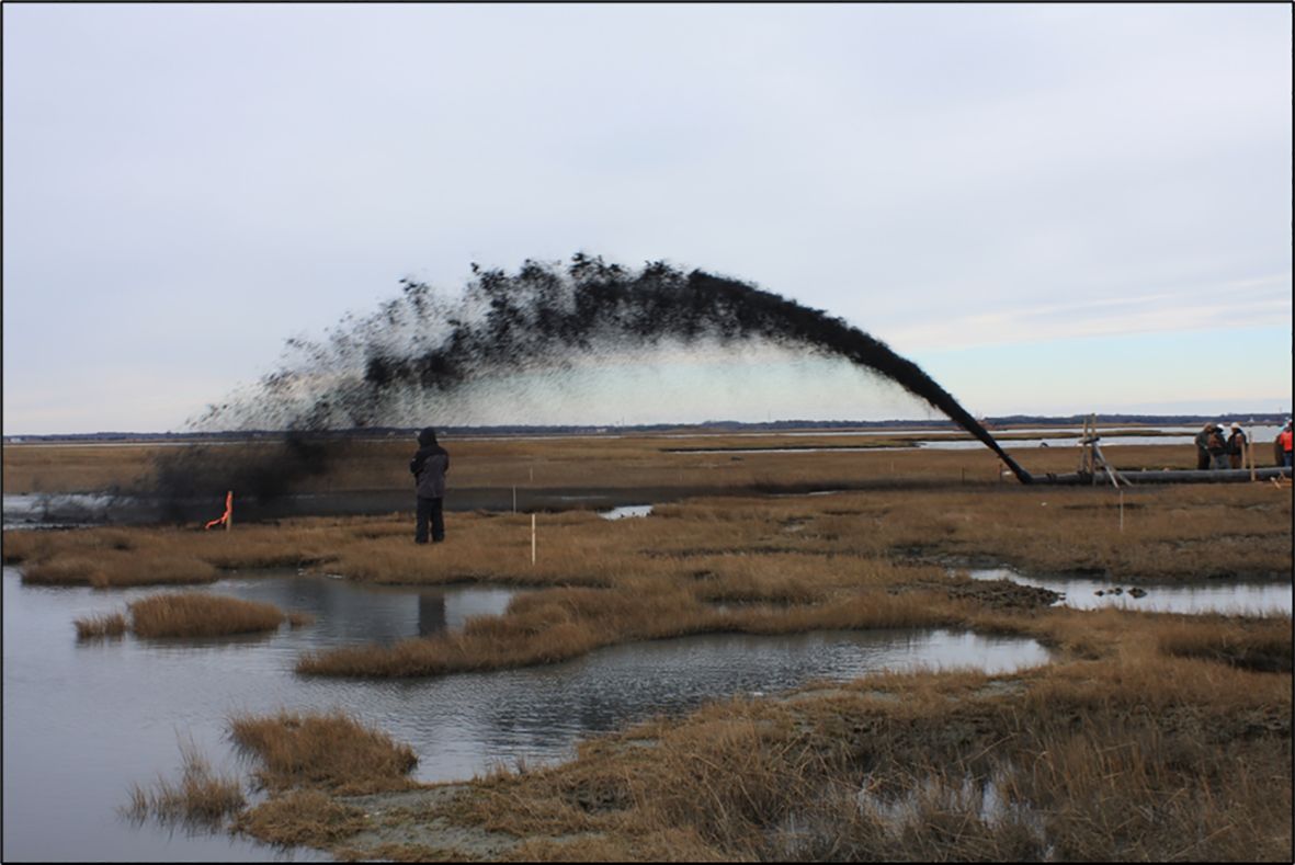 A person stands on a marsh observing a large stream of dark sediment being sprayed into the air from a pipe. The marshland is interspersed with pools of water, and several people are gathered near machinery in the background.