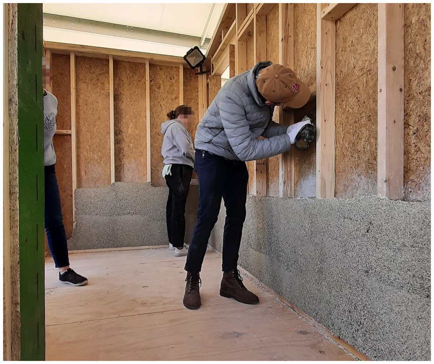 Three students are installing hempcrete into the wall cavities of a prototype structure.