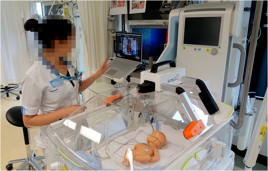 A healthcare professional stands beside a neonatal incubator in a hospital setting, interacting with a nearby computer displaying a video call. The incubator contains a lifelike infant mannequin equipped with monitoring sensors, simulating a medical scenario for training or demonstration purposes.