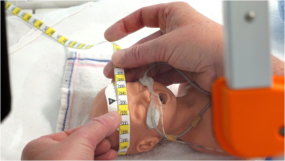 A person measuring the head circumference of a lifelike baby doll with a tape measure. The doll is equipped with a nasal tube and rests on a white cloth.