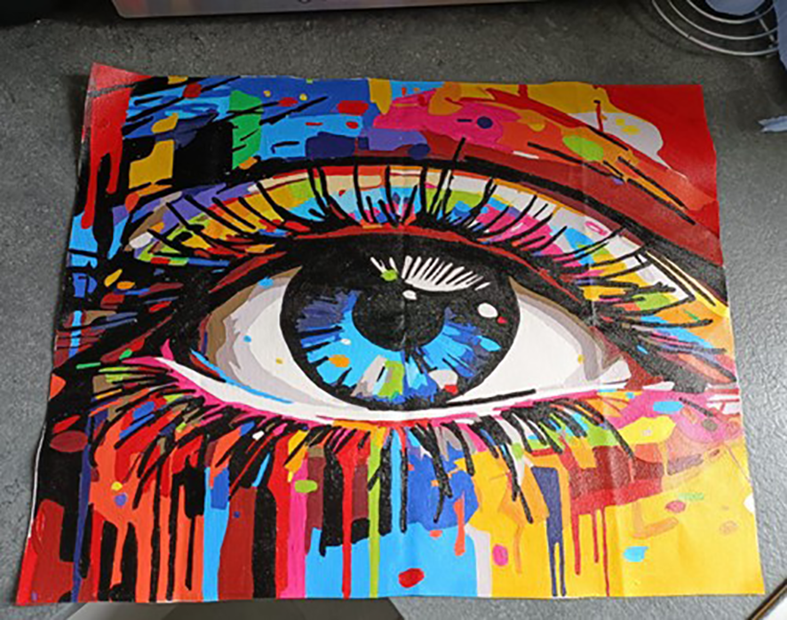 Photograph of a painting on a piece of paper on a counter. The painting is a close up of an eye, made up of squares of multicolour around thick black outlines.