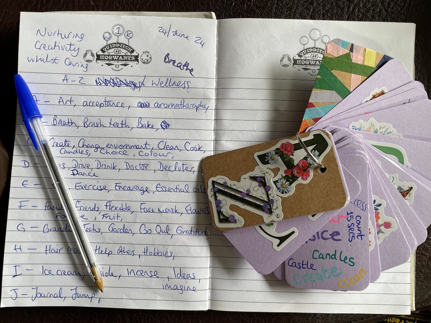Photograph of a lined notebook which is open with a biro on one of the pages. One page is titled ‘nurturing creativity whilst caring’ and the word ‘breathe’ is also written at the top in bold. Handwritten down the lines vertically is the alphabet from A-J, and by each letter are examples (art, acceptance, aromatherapy, breath, brush teeth, bake, create, change environment, clean, cook, candles, choice, colour, drive, drink, doctor, declutter, dance, exercise, encourage, essential oils, friends, family, flexible, face mask, flowers, fruit, garden, go out, gratitude, hair, help others, hobbies, ice cream, incense, ideas, imagine, journal, jump. Sitting over the other page (which is blank) is a key writing with lots of cards on it. The front card has stickers of A and Z on it, and the back has multicoloured washy tape. You can see half of one of the pages which has the examples from ‘c’ on the opposite page in multicoloured handwriting.