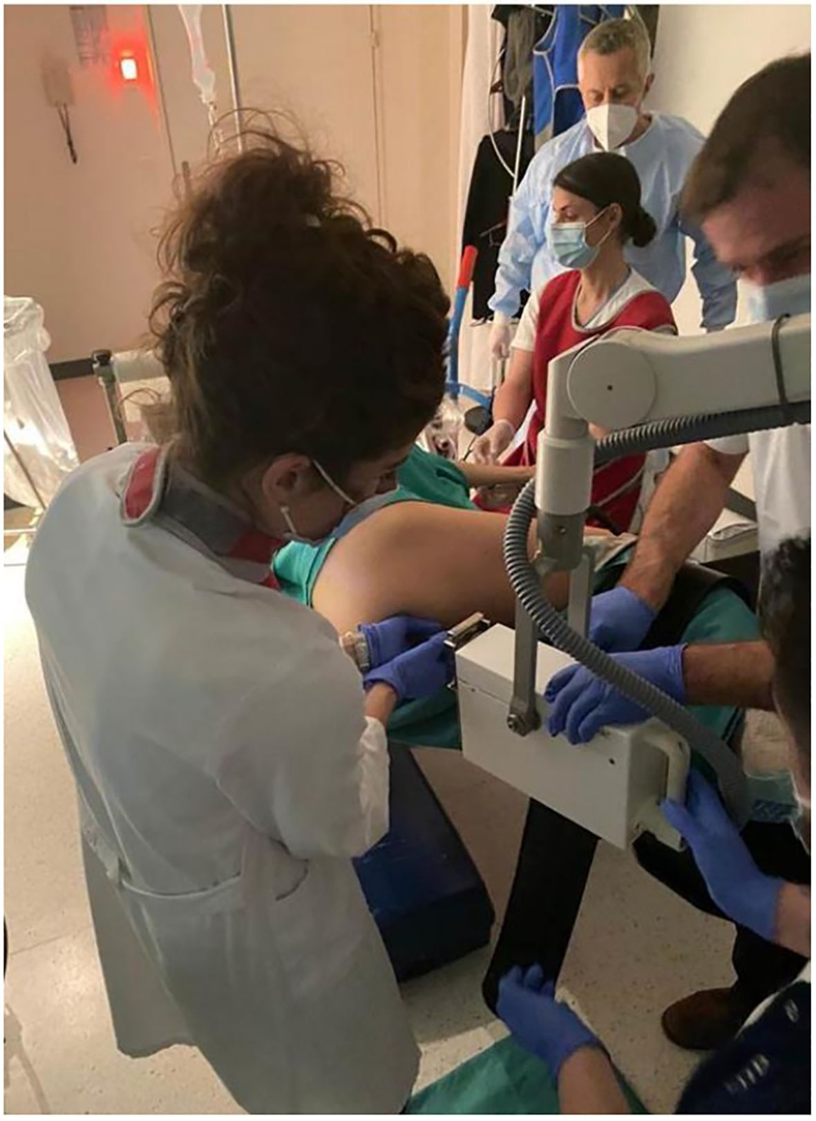Medical professionals wearing masks and gloves are conducting a procedure using a medical device on a person lying face down. The setting appears to be a hospital or clinic room.