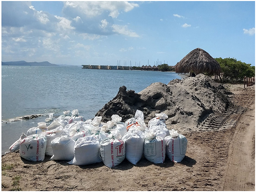 Sandbags are piled on a sandy shoreline beside a mound of earth. In the background, a thatched structure, calm water, and distant hills are visible under a blue sky with clouds.