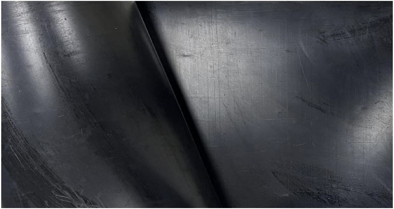 Close-up of a dark metallic surface with a folded edge running diagonally. The surface has visible scratches and reflections, creating a textured and slightly shiny appearance.