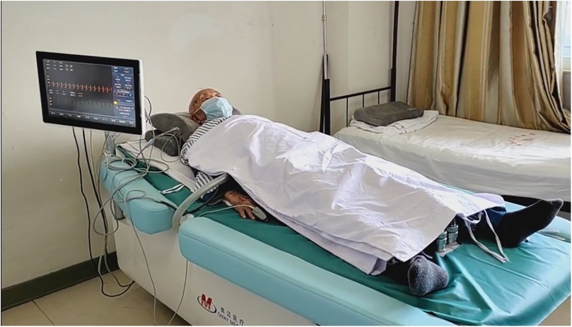 Elderly person wearing a mask rests on a medical bed covered with a white sheet. A monitor displaying vital signs is positioned beside the bed, showing a heart rate graph.
