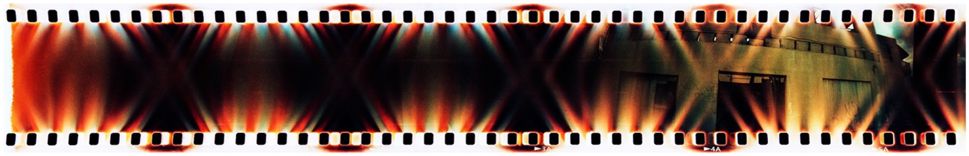 A strip of 35mm film displaying a mix of burnt effects, with colors ranging from dark red to orange and black, creating a dramatic, abstract pattern. The edges are perforated with the conventional sequence of holes found in film strips.