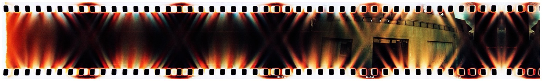 A strip of damaged film showing distortion and multicolored light streaks, predominantly in red and orange hues, with parts of a building faintly visible in the background.