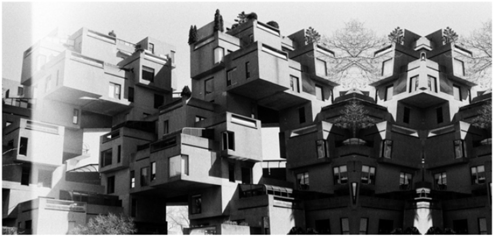 Brutalist architectural structure composed of stacked, modular concrete units resembling cubes. The complex arrangement creates various overhangs and terraces, with minimalistic windows and some foliage seen on rooftops. Monochrome image enhances geometric patterns and contrasts.