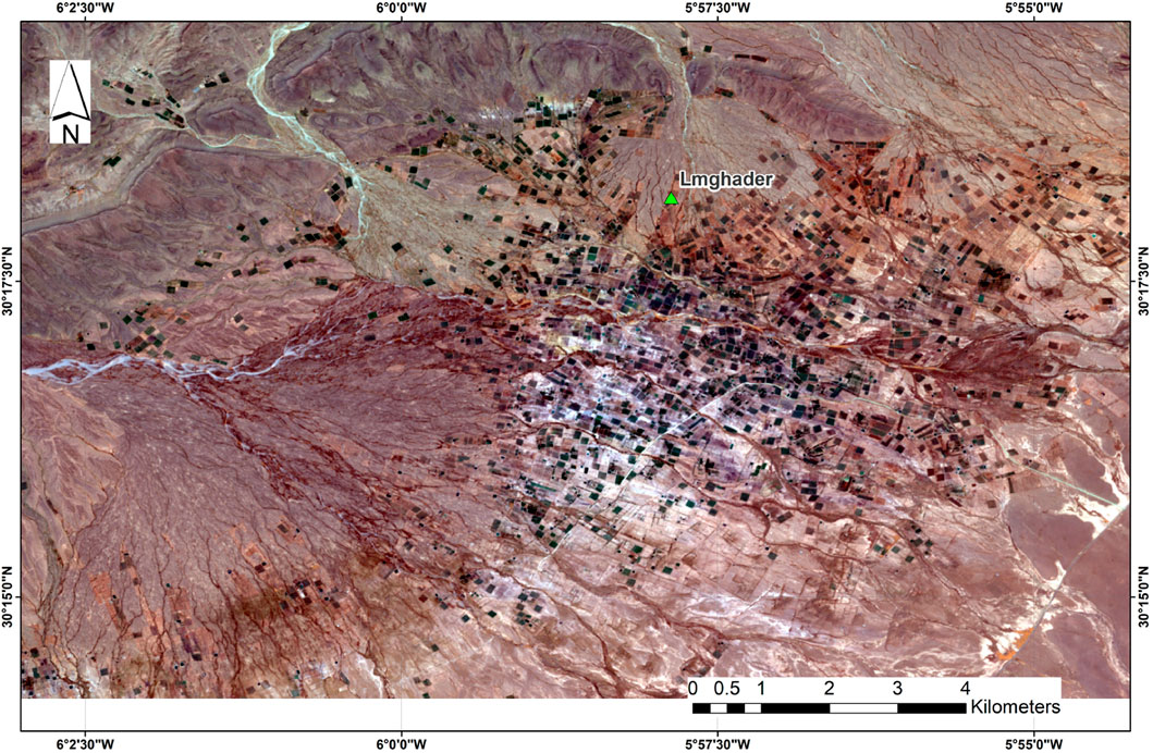 Satellite image showing the area around Lmghader with a desert landscape. Numerous small, dark green agricultural plots are scattered across the terrain. A scale bar at the bottom indicates distances in kilometers, and coordinates are along the edges. A green triangle marks the location of Lmghader.