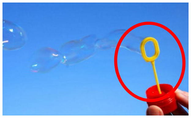 A person holds a bubble wand with a yellow loop at the top, Bubbles float away against a clear blue sky. The bubble wand is highlighted by a red circle.