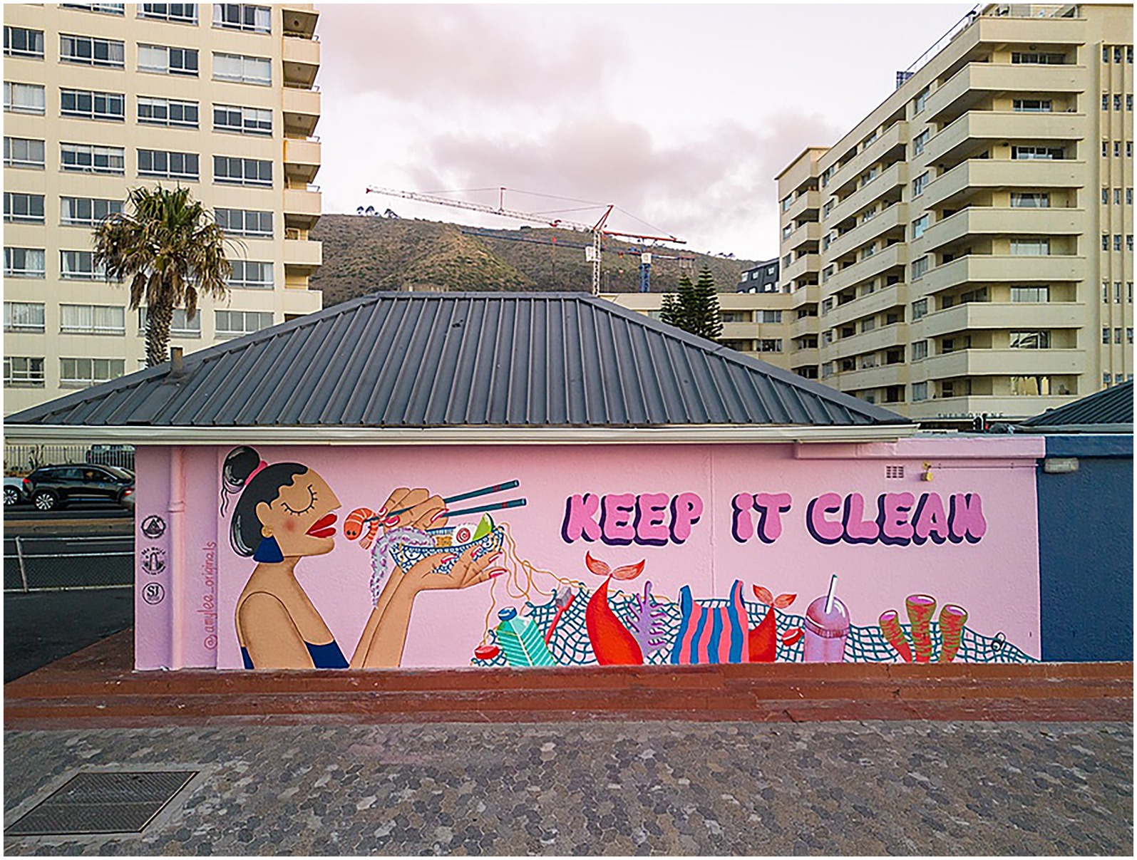 A vibrant mural on a building depicts a woman holding a bowl with chopsticks, eating the seafood that is intermingled with plastic products. Surrounding her are colorful images of fish and trash, accompanied by the message “Keep It Clean” in bold pink letters. The background features tall buildings and a palm tree.