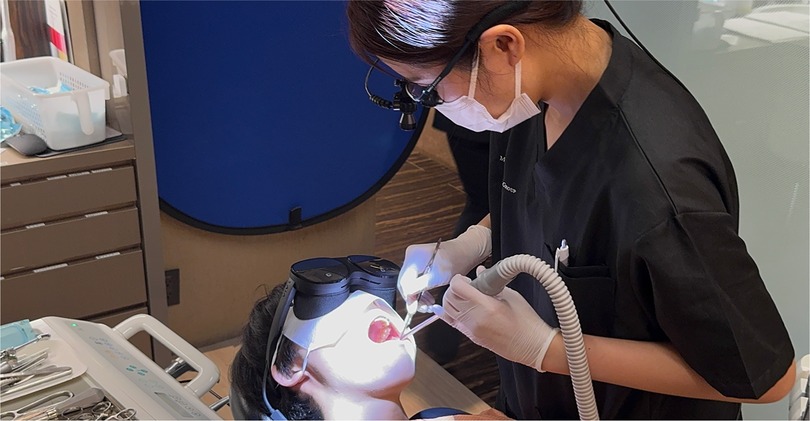 A dental professional wearing magnifying loupes and gloves performs a procedure on a reclining patient using a dental mirror and suction. The patient wears a mixed reality headset, suggesting integration of augmented visual aids during treatment. Surgical tools and equipment are organized on a tray nearby.