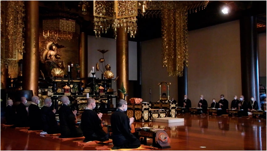 Monks in traditional black robes and masks sit on cushions in a large temple, engaged in a ceremony. The space features ornate golden decorations and a prominent statue at the altar.