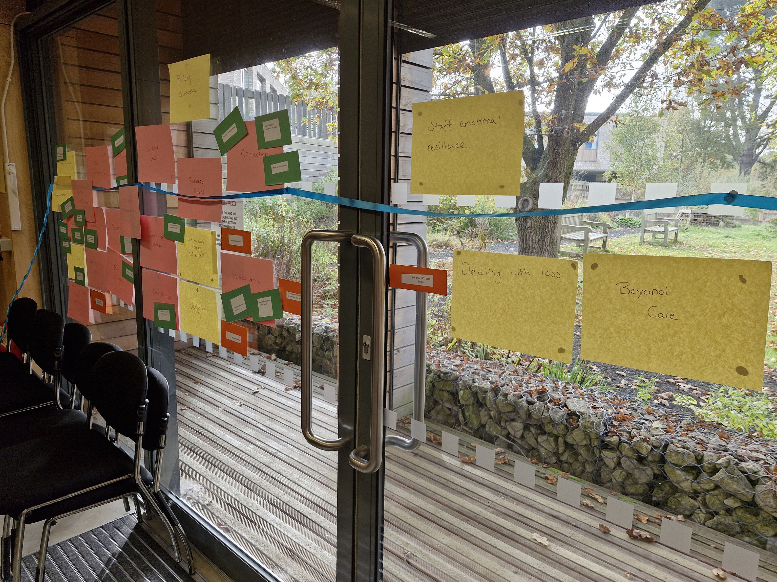 Colored paper notes are taped to a glass door, displaying various topics like “Staff emotional resilience” and “Dealing with loss”. A blue ribbon runs across the door. Outside, a garden with a tree and benches is visible.