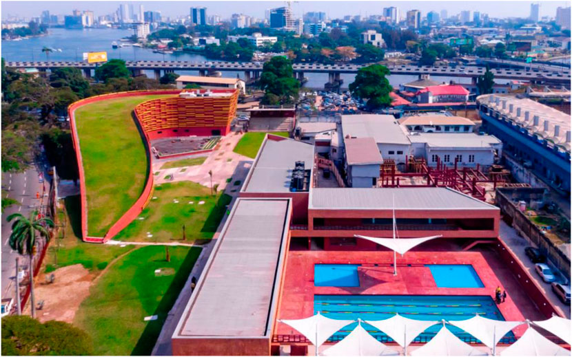Aerial view of a vibrant urban landscape featuring a green-roofed, curved building and a modern complex with a swimming pool. The background showcases a river, bridge, and city skyline.