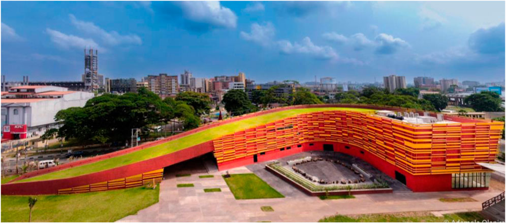 A modern building with a wavy, grass-covered roof, featuring vibrant red and yellow exterior panels. It sits in an urban area with trees and high-rise buildings in the background under a cloudy sky.