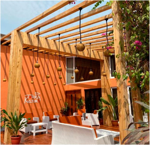 Outdoor seating area with white wicker furniture and orange cushions. A wooden pergola overhead features hanging woven pendant lights. Orange walls and green plants enhance the vibrant setting.
