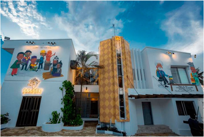 A two-story building exterior with colorful murals depicting people engaged in various activities. The facade includes a large mural with diverse characters and a neon sign reading